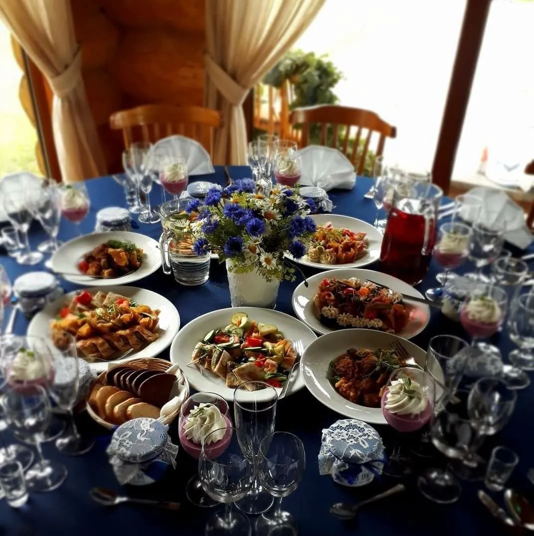 Apaļš klāts galds ar zilu galdautu, rudzupuķu pušķi, ēdieniem un desertiem